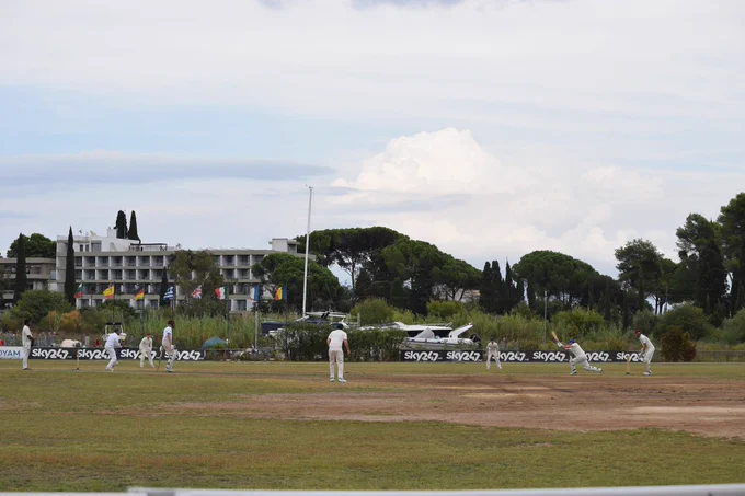View from the boundary in Corfu shows batter hitting ball as fielders and the umpire looks on. Boats, European flags and trees can be seen in the background