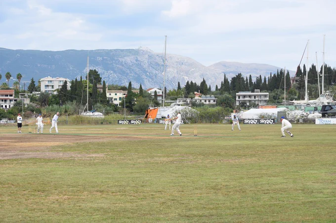 View from the boundary in Corfu shows batter Bobby hitting ball as fielders and the umpire looks on. Boats and mountains can be seen in the background