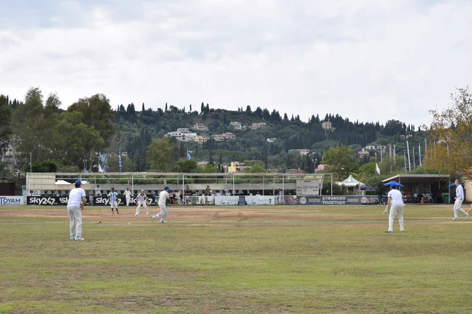 View from the boundary shows batter hitting ball as fielders looks on. Houses and trees under clouds can be seen in the background and a white scoreboard