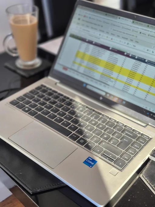 An open laptop on a table, showing a blurred screen of an excel spreadsheet, with a coffee in the background