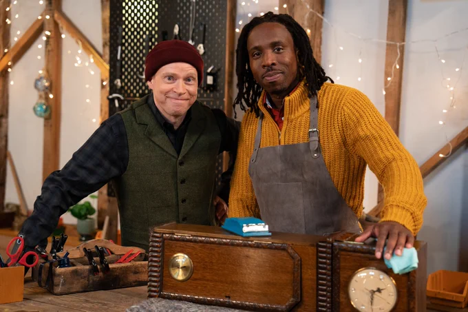 Robert Webb and Kiell Smith-Bynoe wearing warm clothes inside a workshop where a small grandfather clock is being repaired.
