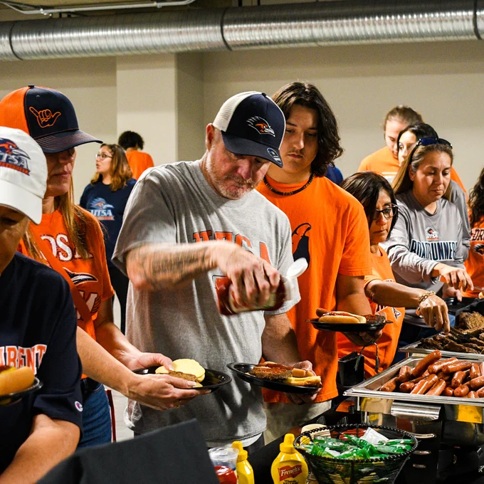 Fans wearing University of Texas at San Antonio apparel serve themselves hot dogs and burgers at a busy indoor tailgate event.