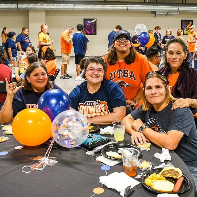 Group of people sitting at a table with food and balloons, wearing University of Texas at San Antonio gear.
