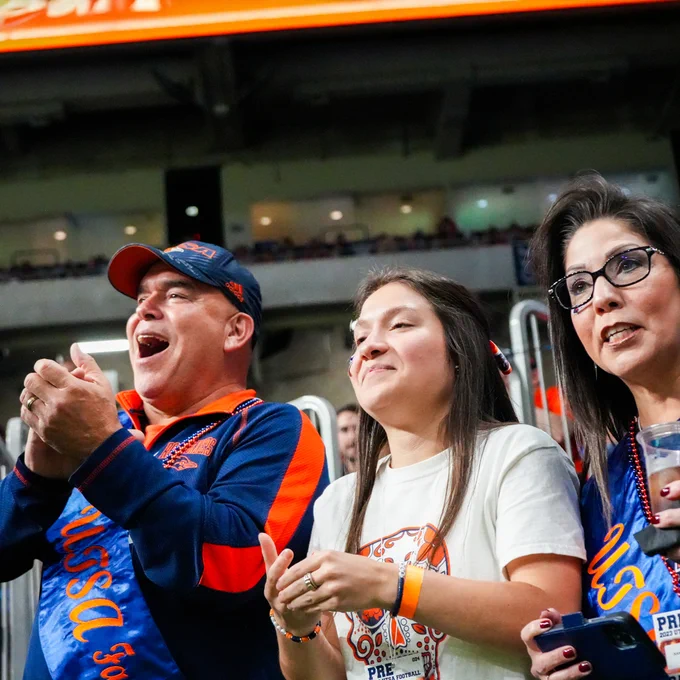 A family of three fans wearing UTSA apparel cheer excitedly at a  football game.