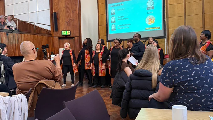 SAWN Choir in black and orange attire performing for seated audience in wood-panelled room with a presentation screen at the 2025 Social Care Future gathering.