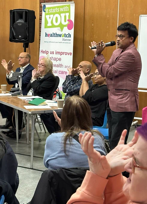 Four panelists sit at a table, with a Healthwatch Harrow banner behind them. A fifth panelist is standing. They are facing an audience. The panelists and audience members are raising their hands. 