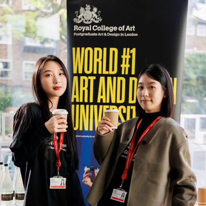 Two people holding a coffee cups, standing in front of a Royal College of Art promotional banner at an event, it read "world #1 art and design university".