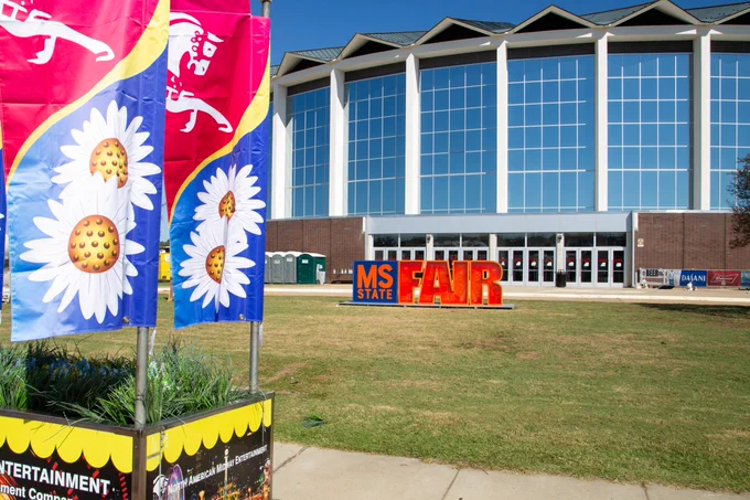 Exterior view of the Mississippi State Fairgrounds with colorful banners in the foreground and a large "MS STATE FAIR" sign in front of the main building.