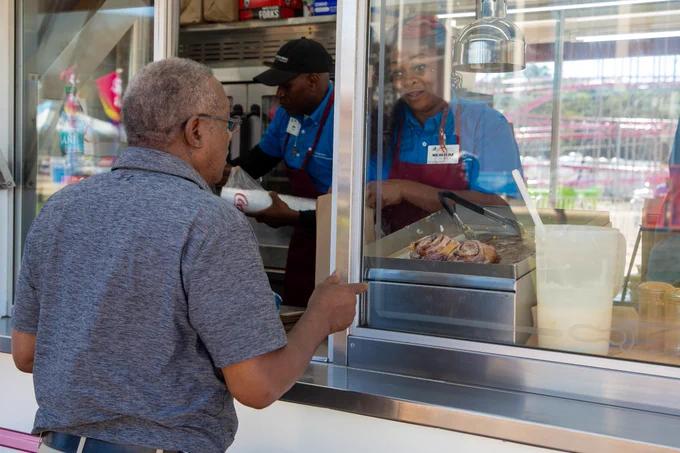 Customer placing an order at a Mississippi State Fair food stand, with workers preparing food and a tray of cinnamon rolls visible in the background.