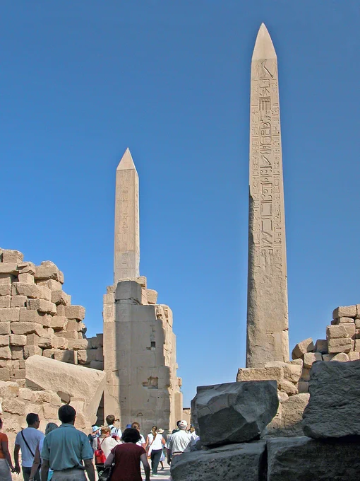 Tourists walk towards two ancient Egyptian obelisks with hieroglyphs, set amidst the ruins of a temple under a clear blue sky.