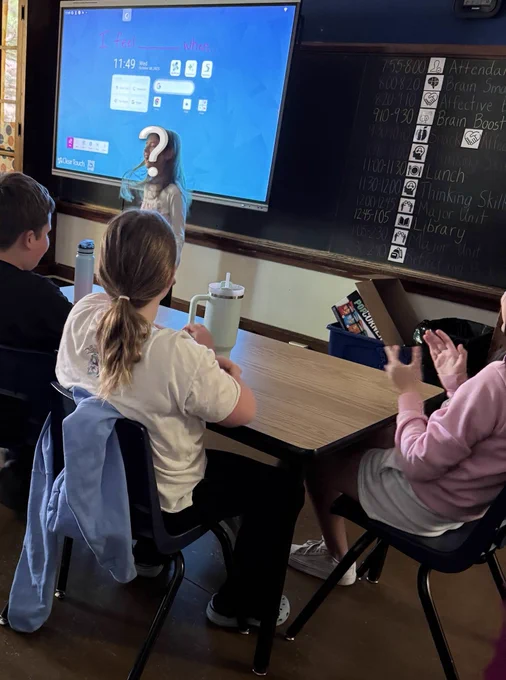 Student standing in front of a screen with a question mark over her face. Students are sitting at tables in front of her. One student is clapping in delight.
