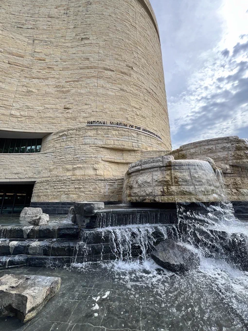 Exterior view of the National Museum of the American Indian, in Washington, D.C., featuring a curved stone facade and a cascading waterfall in the foreground.