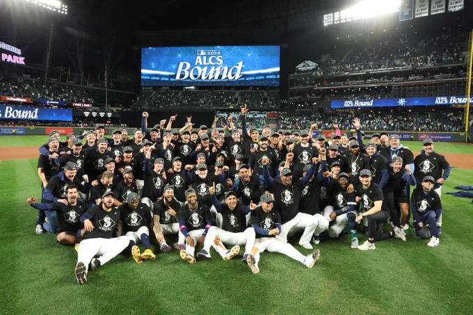 Seattle Mariners players and staff pose for a photo on the infield grass at T-Mobile Park after winning ALDS Game 5 against the Tigers on a 15th-inning walk-off hit. They are wearing black t-shirts with "ALCS 2025" and the Seattle Mariners logo on the front.