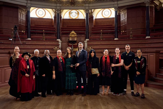 A group of Mākereti's descendants and members of her community, standing a line in an ornate room in front of rows of dark, wood-pannelled seating. Credit: Fisher Studios