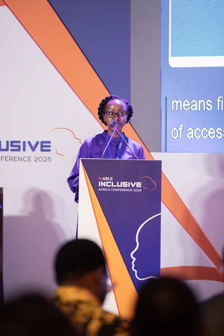 A woman with glasses and braided hair is standing at a podium, speaking into a microphone. The podium is branded with the words "inABLE INCLUSIVE AFRICA CONFERENCE 2025." The background is a white and orange graphic. Part of a screen is visible to the right with the words "means fi..." and "of access...". In the foreground, the backs of people's heads are blurred.