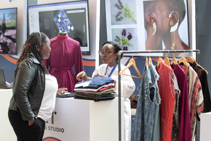 Two people are standing at a white exhibition booth labeled "Hisi Studio." A woman with braids and a black leather jacket is talking to another woman in a white blouse and glasses, who is showing her a stack of folded clothes. Behind them, clothes hang on a rack and a large screen shows a computer desktop.