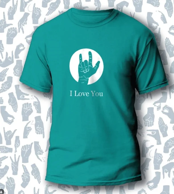 A teal-colored T-shirt with a large white circle on the front. Inside the circle, a hand shows the American Sign Language sign for "I love you." The words "I Love You" are printed below the circle. The background is a pattern of various hand signs in shades of gray.