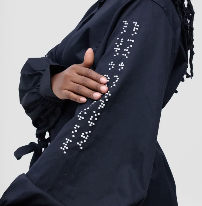 A close-up shot of a person's arm and torso. They are wearing a black jacket with a pattern of raised white dots down the sleeve, which is Braille. Their other hand is resting on their chest, with a black belt tied around the waist.