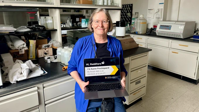 Karen Prestegaard wearing a blue shirt and glasses, holding a laptop in her lab to promote her Reddit Ask-Me-Anything on September 29, 2025