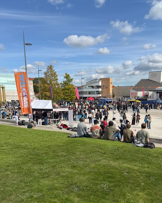 Students gather outdoors at the University of Warwick Piazza with tents, banners, and a clear blue sky overhead.