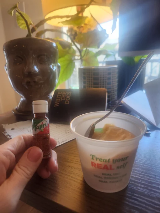 I'm holding up a tiny bottle of Tajin seasoning next to a frozen mango cup on my desk, which has some papers and plants on it as well.
