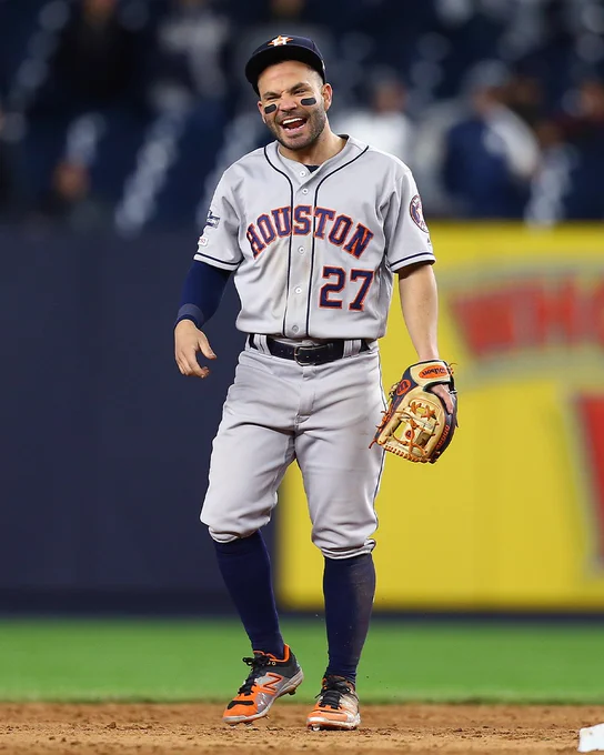 Jose Altuve stands in the infield at Yankee Stadium and laughs. He is wearing a gray road Astros uniform with blue lettering and orange trim and dark blue Astros cap.
