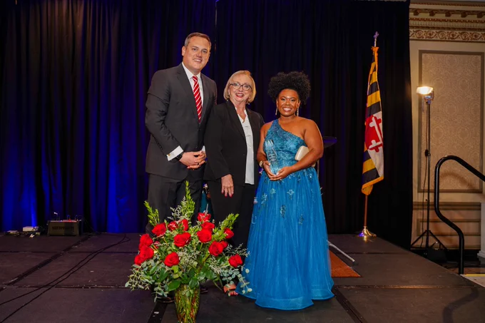 Three people pose at an award ceremony, flanked by red roses and a Maryland flag in the background.