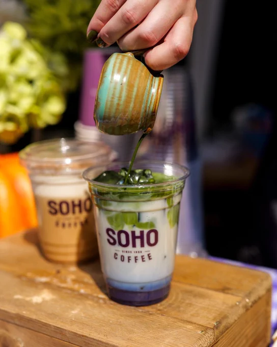 A person is pouring a green matcha layer from a small jug into a cup of iced milk branded with "SOHO COFFEE". The beverage is on a wooden counter beside another similar drink.