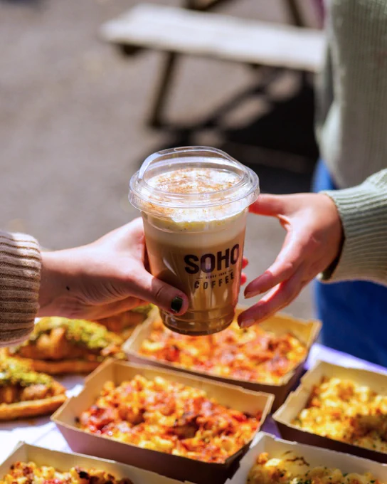 Two individuals exchanging a cup of SOHO Coffee, with boxes of colourful food in the foreground.