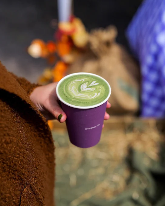 A person holding a cup of matcha latte with a beautiful leaf design on top, against a blurred background of straw and autumn decorations.