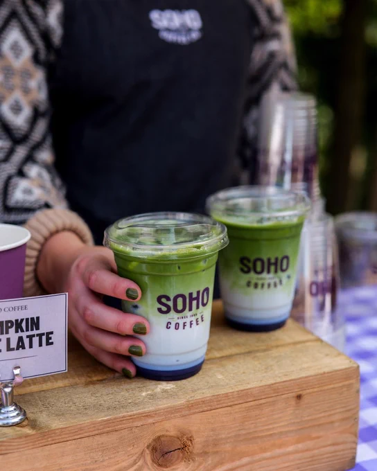 A person holding a tray with two cups of matcha latte from SOHO Coffee. at an outdoor event.