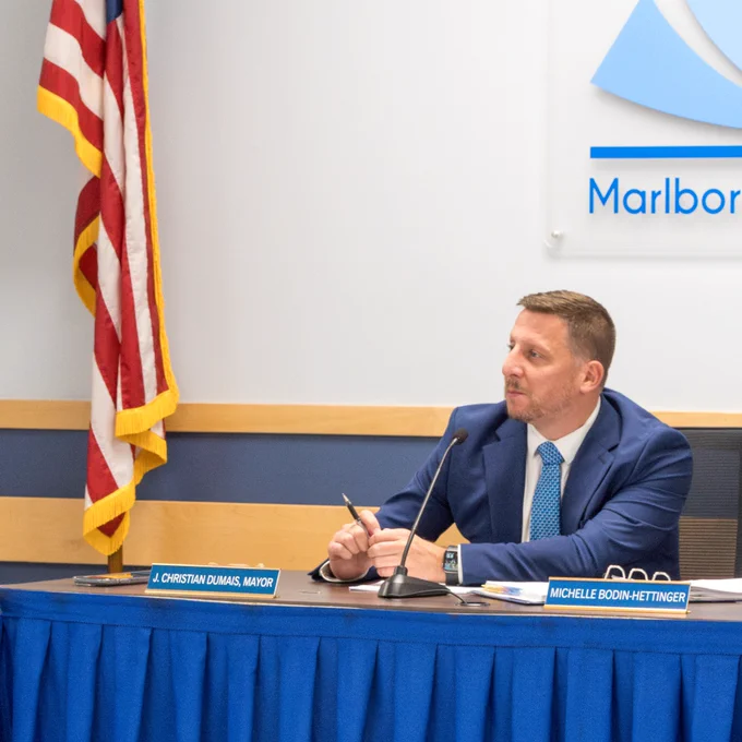 Marlborough School Committee Chair, Mayor Christian, sits at a table with the USA flag to his right and the MPS logo behind him. 