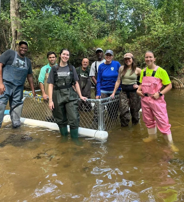Eight volunteers stand in a stream wearing waders and holding a trash trap. Photo credit: North Carolina Sea Grant
