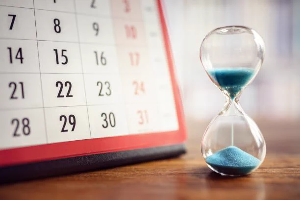 Close-up of a calendar showing dates and an hourglass with blue sand running through it, sitting on a wooden desk.