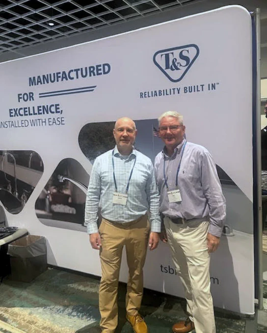 Two individuals standing in front of a T&S Brass promotional banner at the ASPE Symposium. Both are smiling and wearing business casual attire with event lanyards.