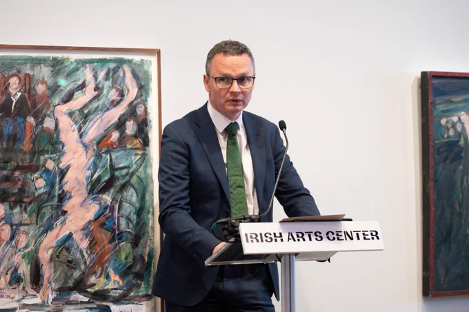 Patrick O’Donovan TD, Ireland’s Minister for Culture,
Communications and Sport, speaks from a dais on which there is a plaque that reads, Irish Arts Center. Two oil paintings hang in the background.