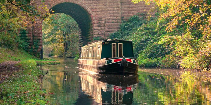 A dark green narrow boat with red and gold details cruises along the canal. A brick bridge with an arch is in the background, and both sides of the canal are lined with autumnal foliage showing shades of green and orange.