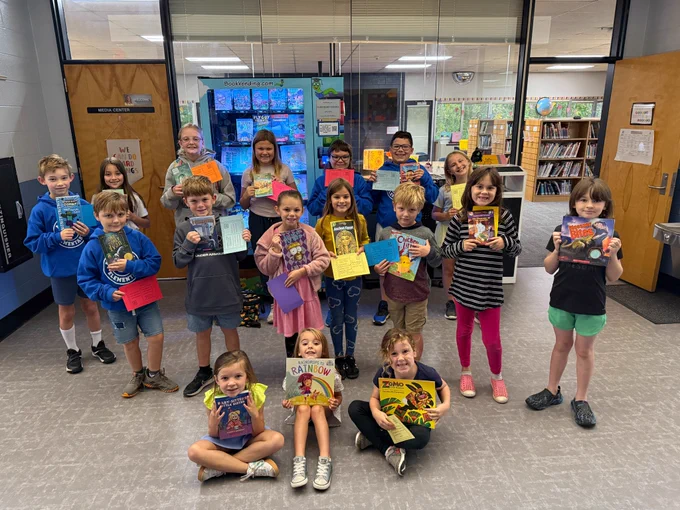 17 students gather around the book vending machine outside of the library. they are all smiling and holding their books and certificates