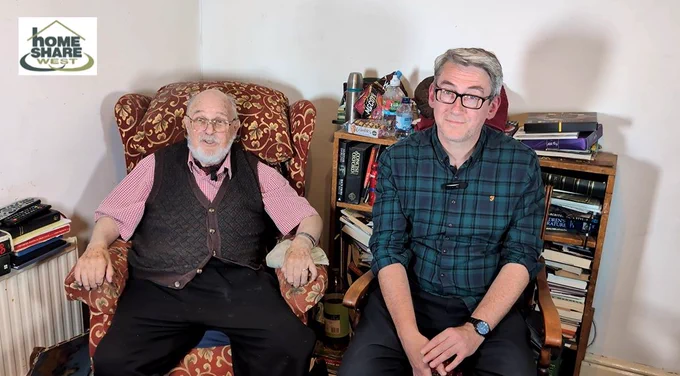 Alt text for image: Two men sit together in a cosy living room filled with books. The older man on the left has white hair and a beard, wearing glasses, a red checked shirt and dark waistcoat. The younger man on the right has grey hair, glasses and a green checked shirt. A ‘Homeshare West’ logo appears in the top-left corner.