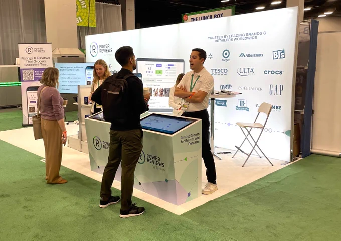 A PowerReviews team member in a white polo shirt speaks with an attendee at the Groceryshop booth. They are standing at an interactive kiosk, and behind them, a large, brightly lit wall displays the logos of well-known brands and retailers like Target, Crocs, and Estée Lauder under the heading "Trusted by Leading Brands & Retailers Worldwide."