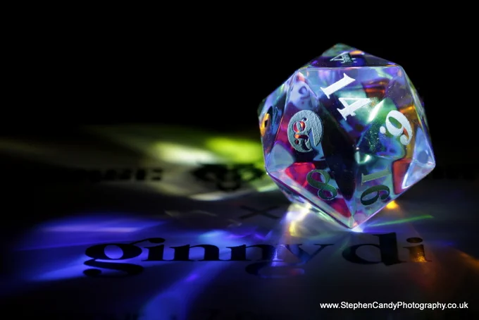 Glass d20 die refracting light in different colours, stood on its box with the Ginny Di and Norse Foundry logos.