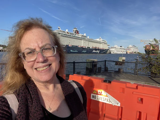 A smiling Lisa (Caucasian woman beyond middle age with long dark hair) across the channel from a TUI ship, a Viking Cruises ship, and a Norwegian Cruise Line ship