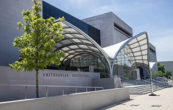 Exterior view of the National Air and Space Museum with a "Smithsonian Institution" sign out front. The building features modern architecture with a curving metallic canopy and large glass panels under a clear sky.