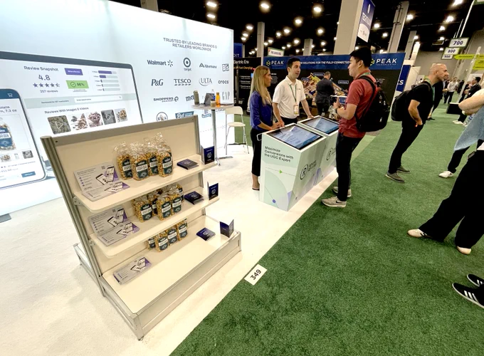 A wide shot of the PowerReviews booth at Groceryshop. On the left, a display stand holds bags of popcorn from Sunrise Snack Co. for the giveaway. In the center, a PowerReviews team member in a white polo shirt speaks with two attendees at an interactive kiosk. The back wall of the booth features a large graphic showing client logos like Walmart, Target, and Tesco, along with a mock-up of the PowerReviews platform interface on a laptop.