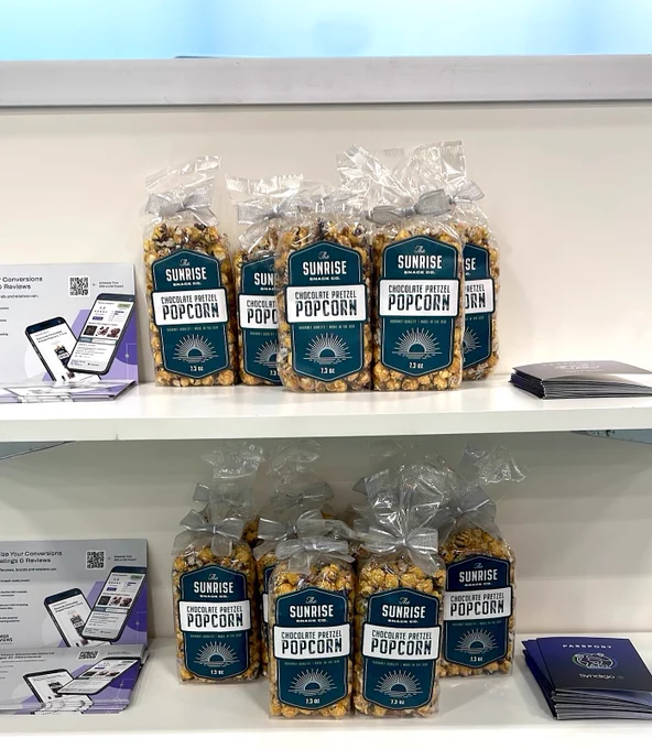 A close-up of a white two-tiered display shelf at the Groceryshop booth. Both shelves are neatly stocked with clear bags of "Sunrise Snack Co. Chocolate Pretzel Popcorn," which are tied with silver ribbons. Next to the popcorn are stacks of promotional flyers, and on the bottom shelf, dark blue "Passport by Syndigo" booklets are also visible.