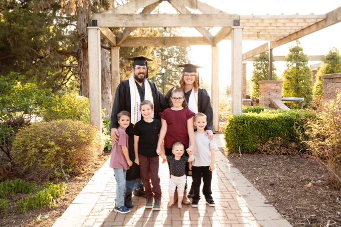 One male and one female college graduates stand with 5 children in an outdoor garden setting. 