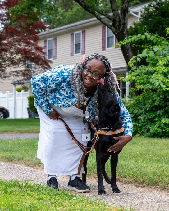 Katina is outside on a sidewalk, bending down and hugging her black Labrador guide dog, Amber, who is wearing a harness. 