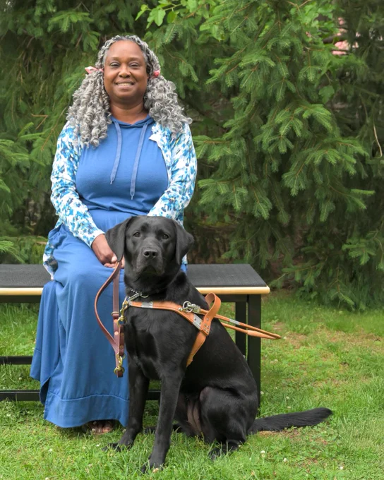 Katina is sitting on a bench outdoors, smiling, with Amber sitting in front of her on the grass. Amber is wearing her harness and looking toward the camera.
