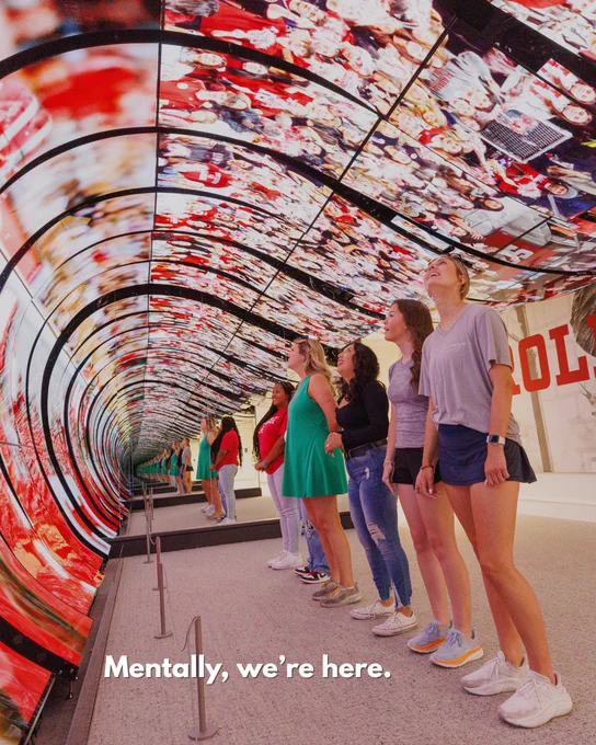 Visitors viewing a large, cylindrical multimedia installation with numerous images displayed, located at the Randall Welcome Center. The words "Mentally, we're here." are on the screen.