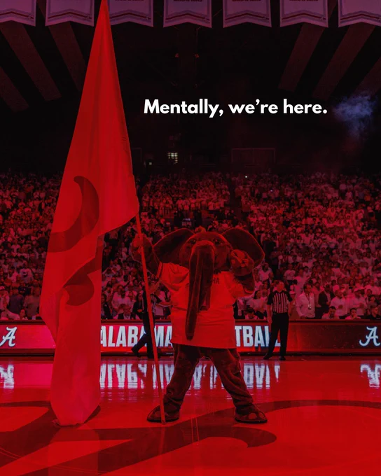 Mascot of the University of Alabama, Big Al, holds a flag with a large Script A at a basketball game with the crowd in the background and the text 'Mentally, we're here.'